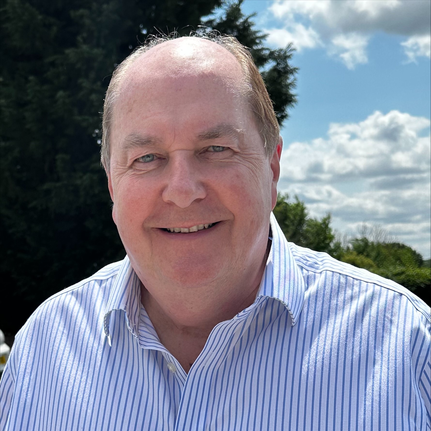 Smiling man in a striped shirt standing outdoors on a sunny day with trees and clouds in the background.