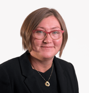 Woman with short brown hair, red glasses, black blazer, and gold necklace, smiling at the camera.