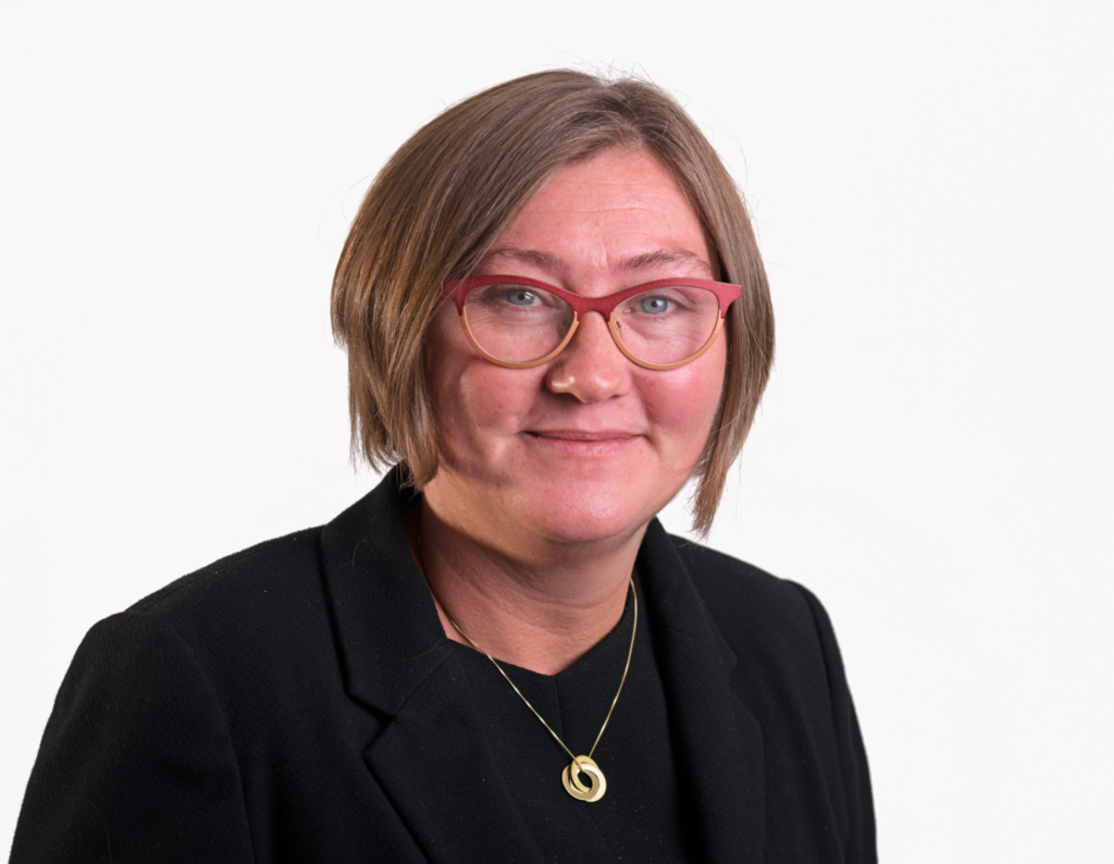 Woman with short brown hair, red glasses, black blazer, and gold necklace, smiling at the camera.
