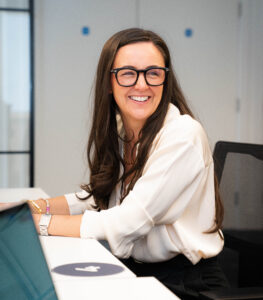 Woman with long dark hair and glasses smiling at a desk in an office setting.
