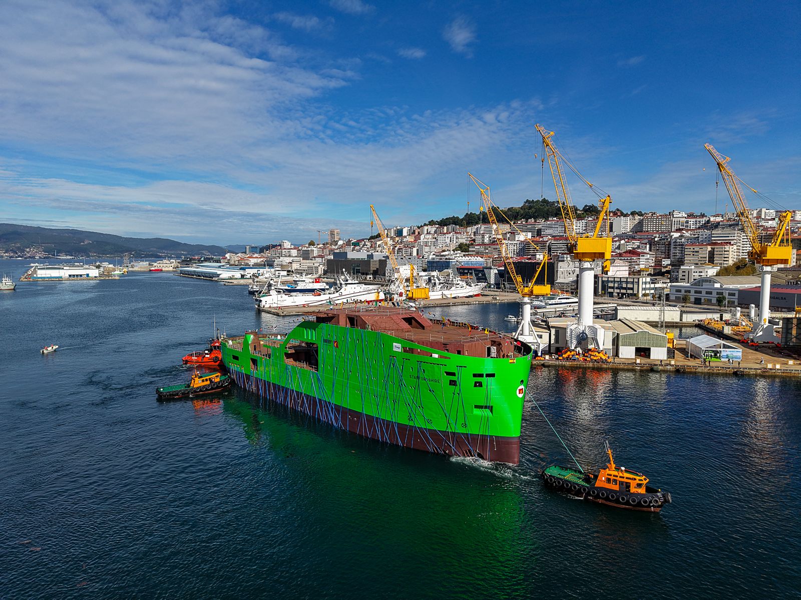 Two tugboats manoeuvring a large, bright green ship hull in a harbour.