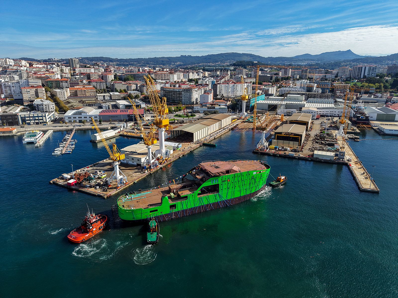 Tugboats manoeuvre a large, bright green ship hull in a shipyard harbour.