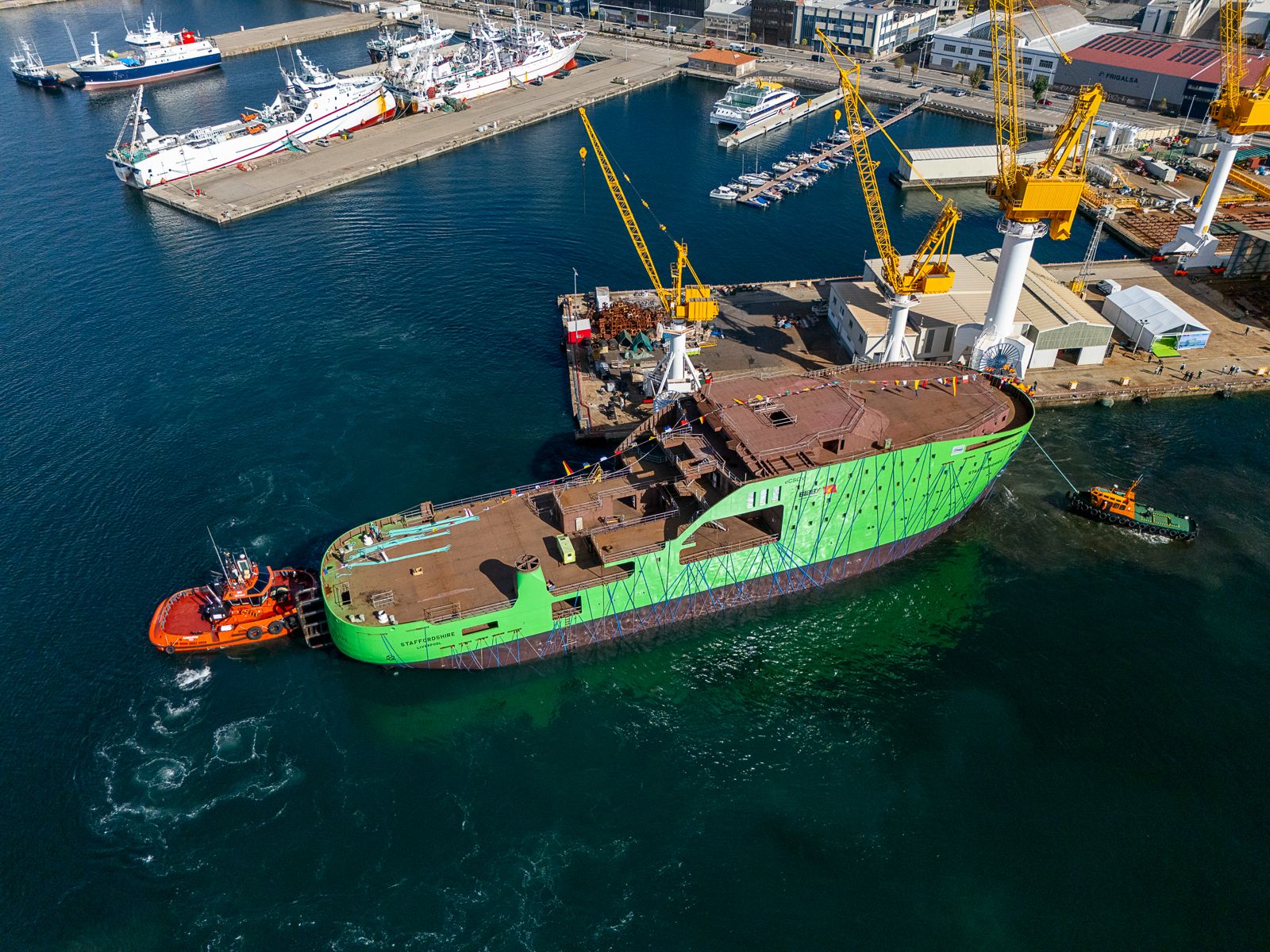 A large green ship hull being manoeuvred by a tugboat in a harbour.