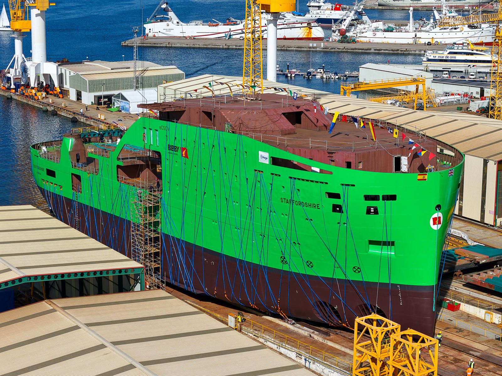 A large green and brown ship hull under construction at a shipyard.