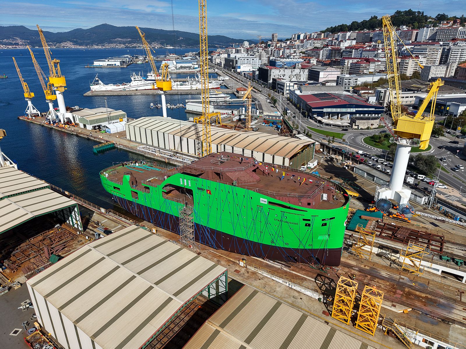 A large green ship hull under construction in a shipyard beside a city harbour.